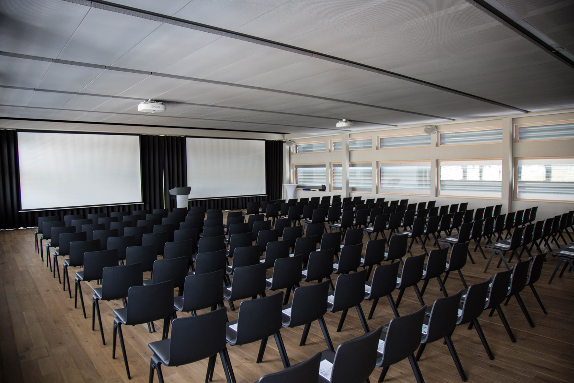 Konferenzraum mit Reihen schwarzer Stühle in Theateranordnung vor zwei großen Projektionsleinwänden und einem Podium, mit Holzfußboden, Deckenbeleuchtung und Fenstern mit Jalousien an der Seitenwand.