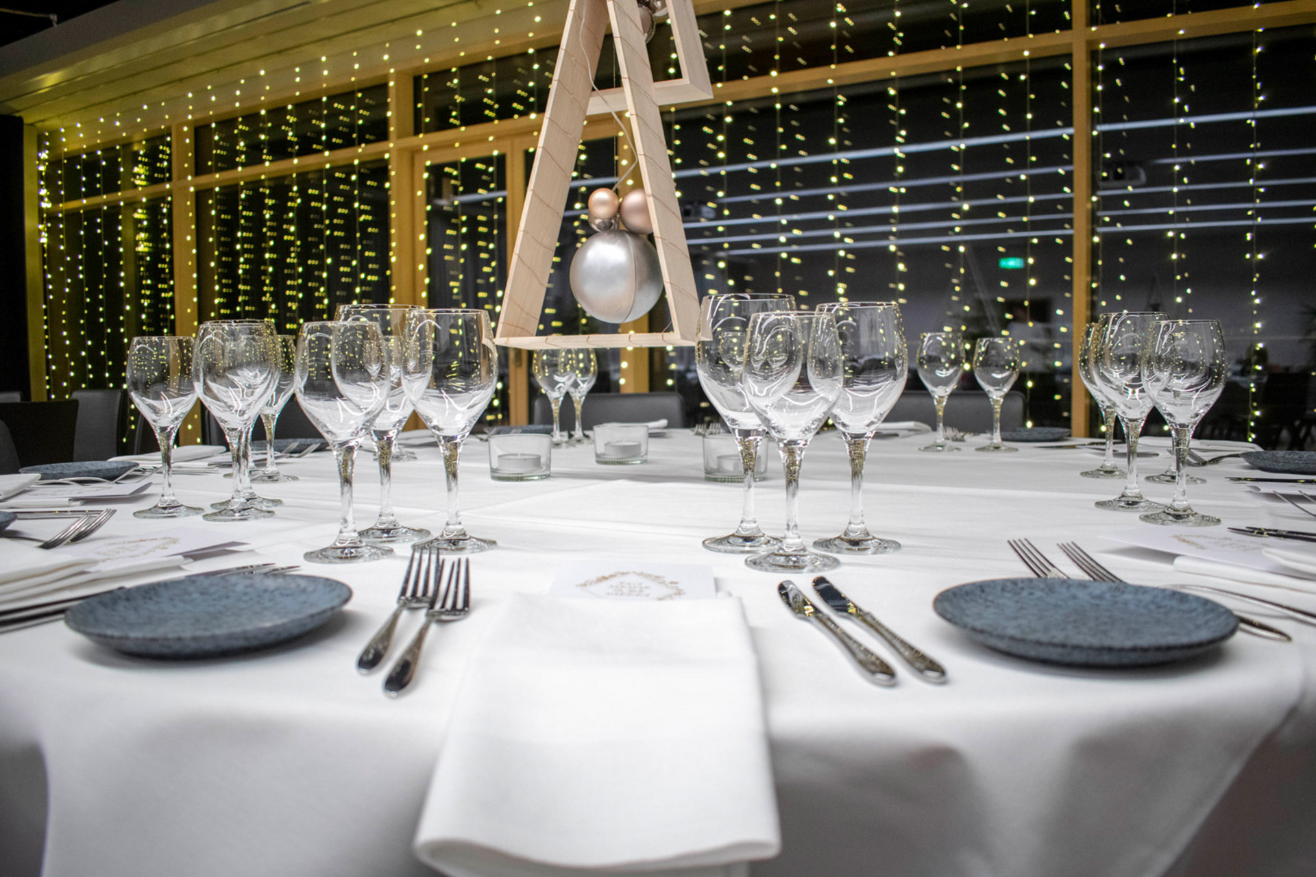 Elegant gedeckter runder Tisch mit weißer Tischdecke, mehreren Weingläsern und festlichem Mittelstück vor einer Kulisse aus Lichterketten.