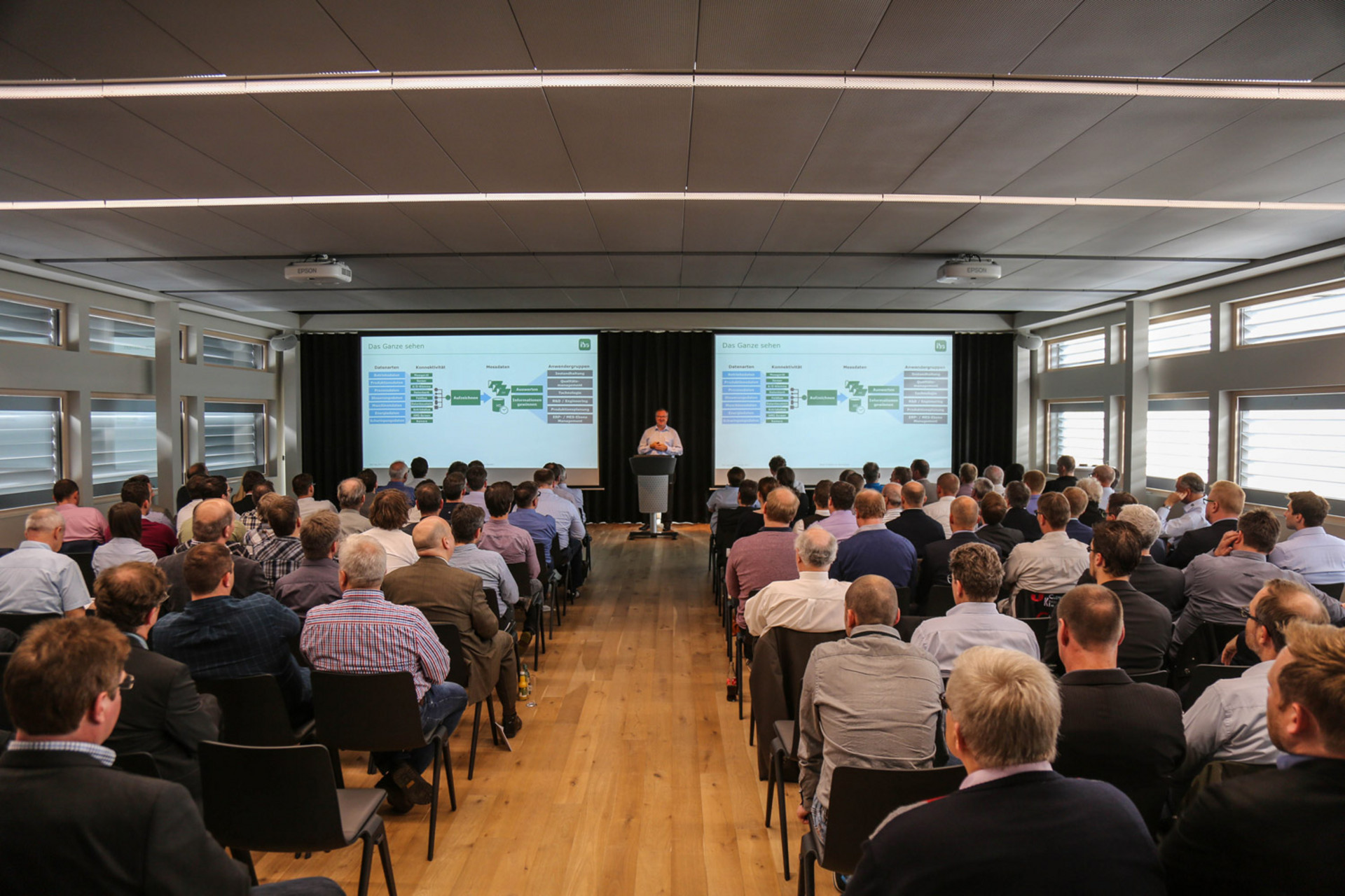 Konferenzraum mit Teilnehmern, die in Reihen sitzen und auf ein Podium sowie zwei große Projektionsleinwände blicken, in einem modernen Raum mit Holzfußboden, Deckenbeleuchtung und breiten Fenstern an den Seitenwänden.
