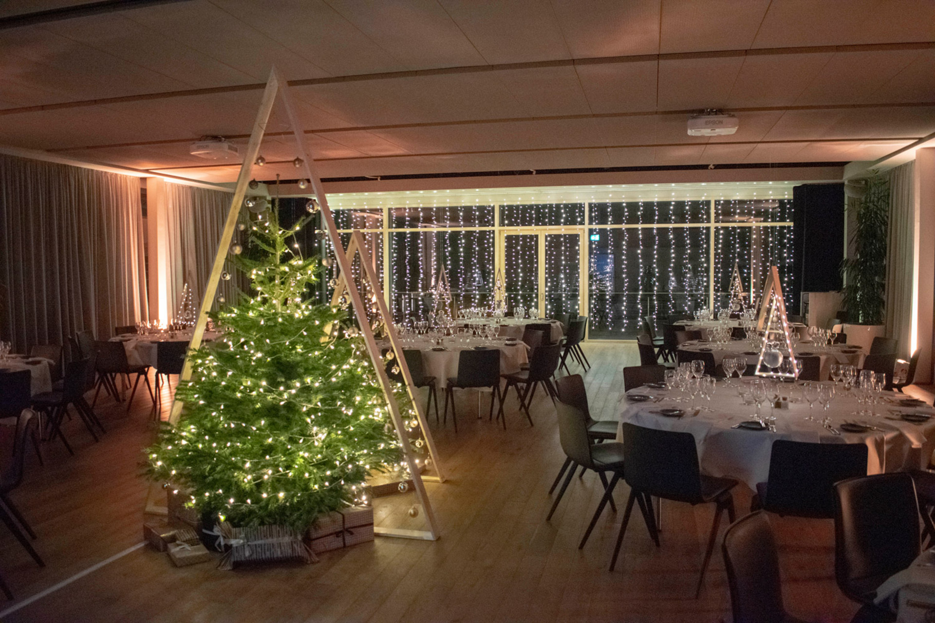 Festlicher Speisesaal mit runden Tischen, weißen Tischdecken, Gläsern und Besteck, geschmückt mit beleuchteten Weihnachtsbäumen und Lichterketten.
