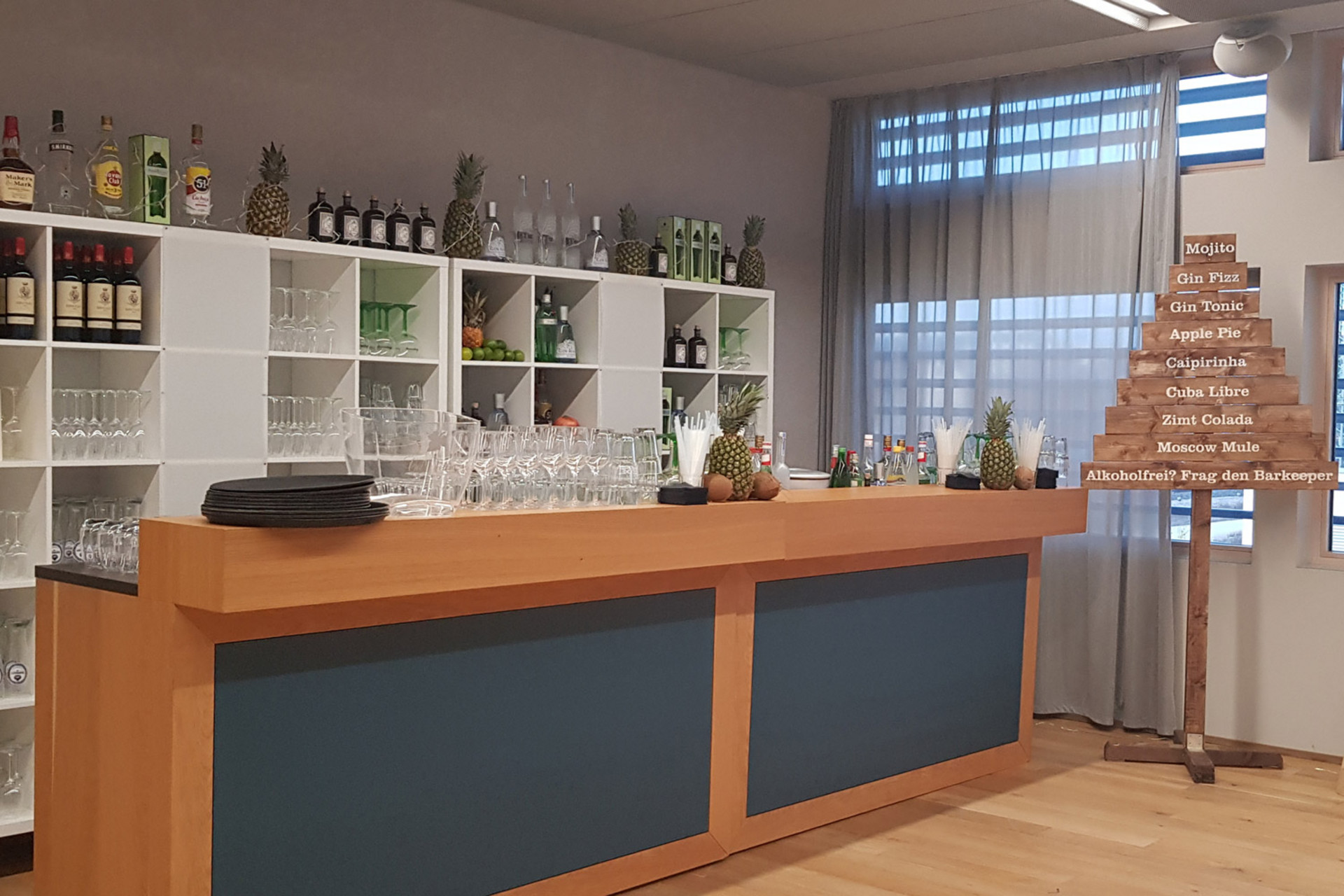 Moderne Bar mit Holztresen, Gläsern, Spirituosenflaschen, frischen Ananas und einem Holzschild mit Cocktailliste.