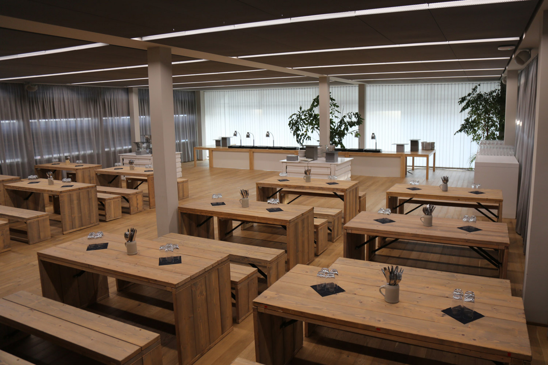 Geräumiger Innenbereich mit langen Holztischen und Bänken in Reihen, gedeckt mit Besteck und Servietten, im Hintergrund ein Buffetbereich sowie große Fenster mit Vorhängen und Topfpflanzen.