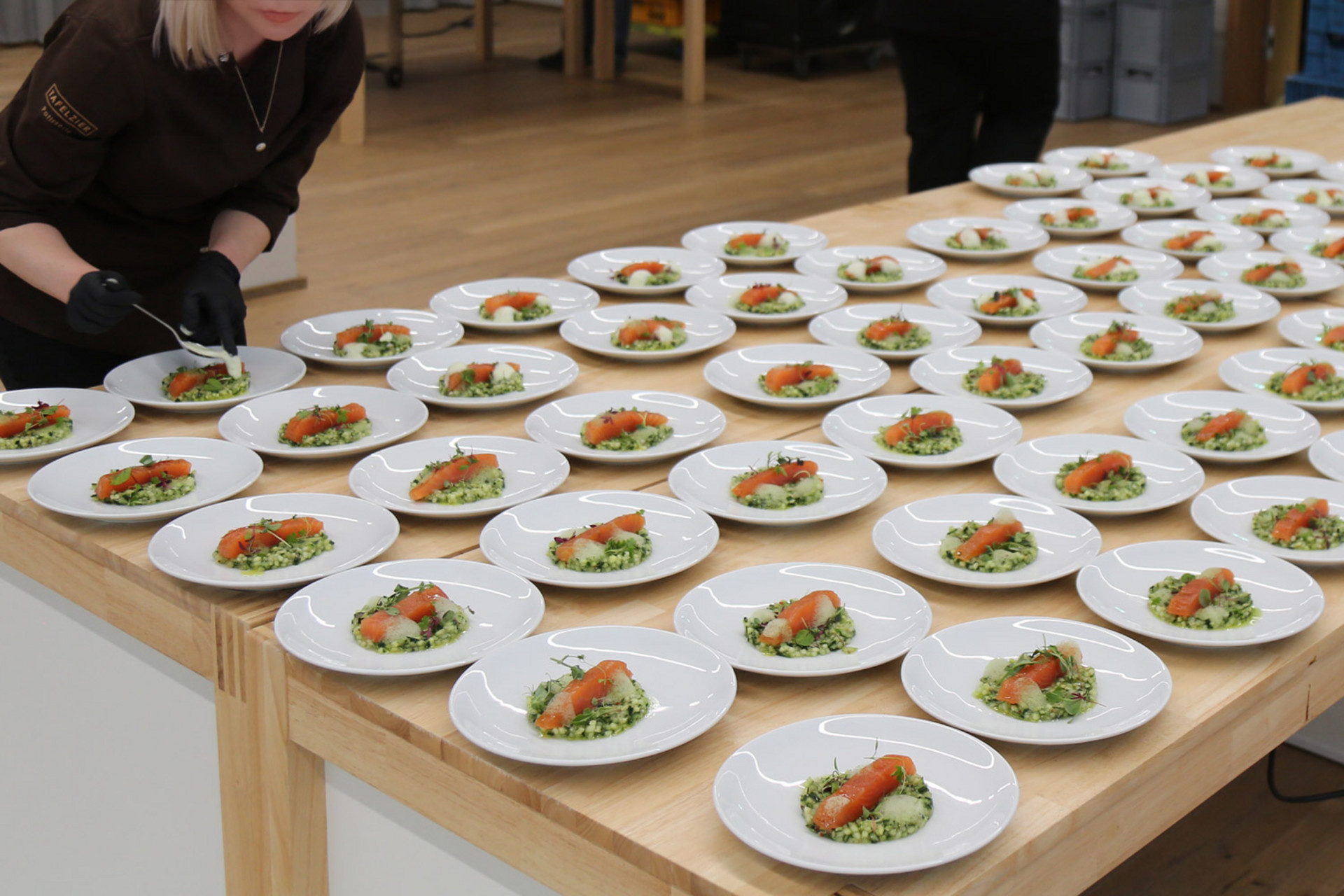 Viele weiße Teller mit Vorspeisen aus Lachs und Kräutern auf einem großen Holztisch, während ein Caterer die Gerichte anrichtet.