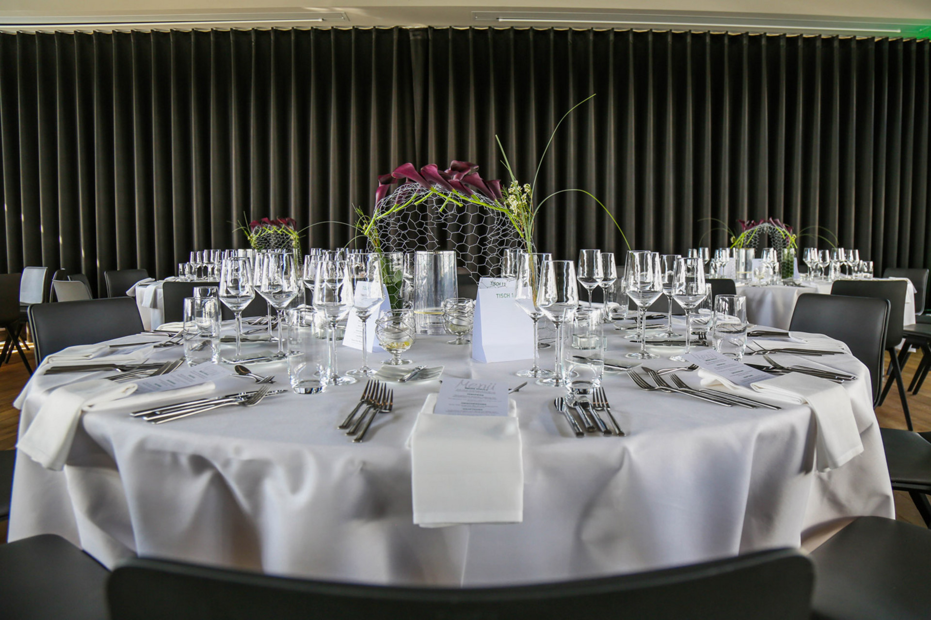 Runder Tisch elegant gedeckt mit weißer Tischdecke, Gläsern, Besteck und einem dekorativen Arrangement aus dunkelvioletten Blumen.