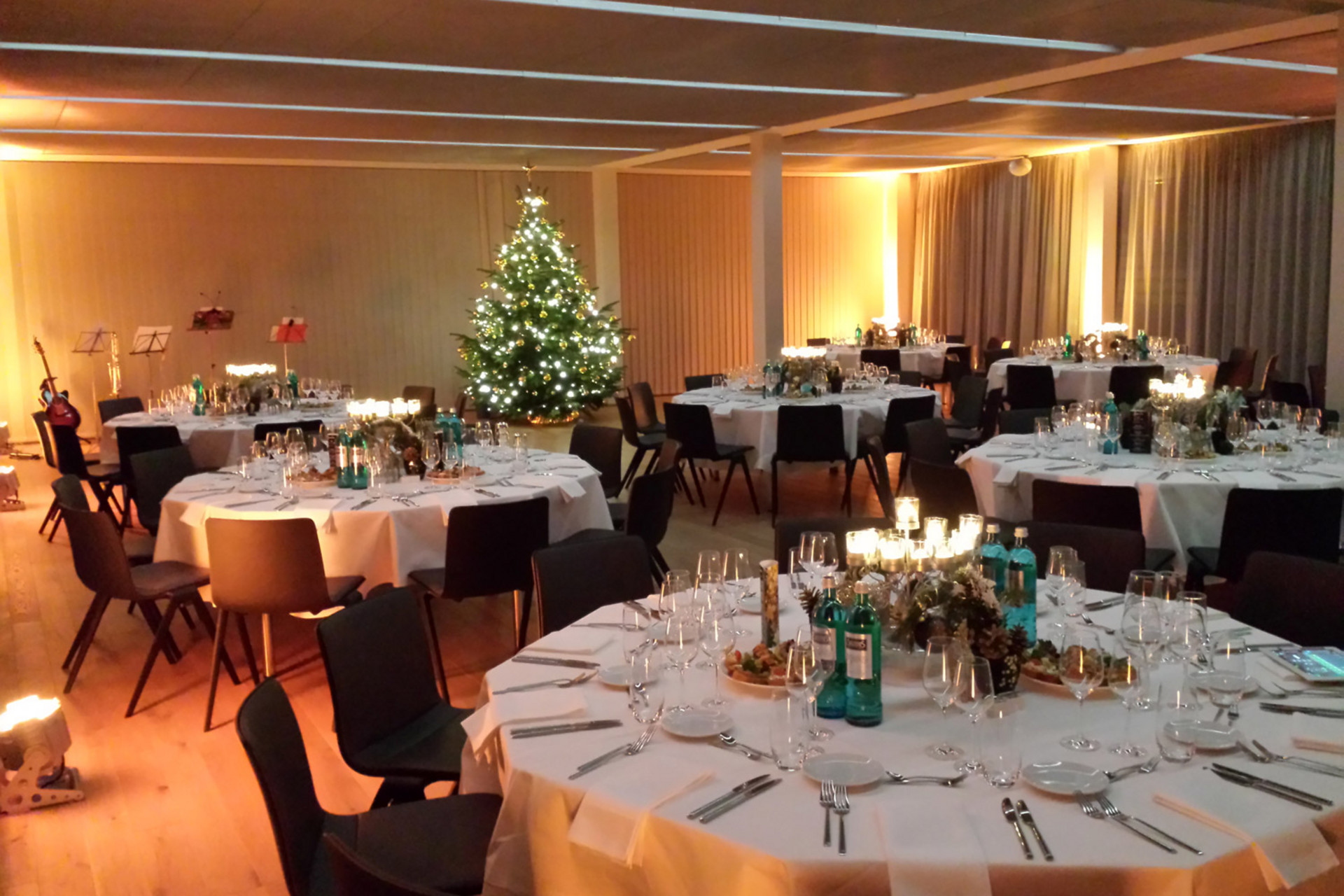 Festlich dekorierter Veranstaltungsraum mit runden Tischen, Kerzen und einem beleuchteten Weihnachtsbaum.
