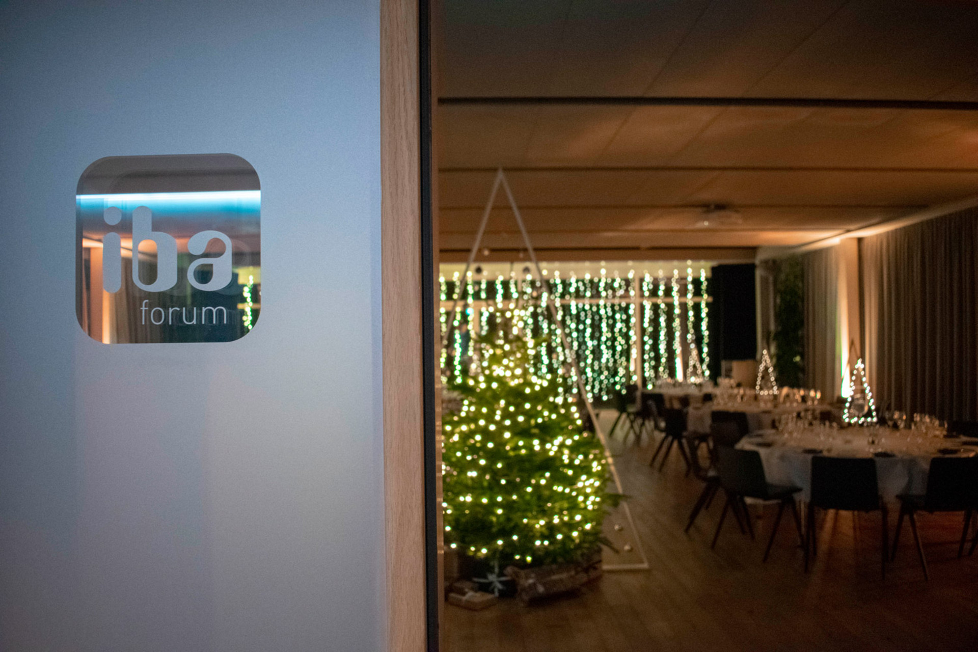 Nahaufnahme einer Glastür mit dem Logo „iba forum“, dahinter ein festlich dekorierter Eventsaal mit beleuchteten Weihnachtsbäumen, Lichterketten und runden, gedeckten Tischen.