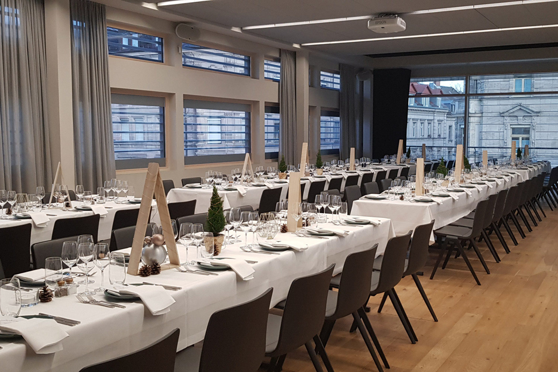 Lange festlich gedeckte Tafeln mit weißen Tischdecken und eleganten Gläsern in einem modernen Eventraum.