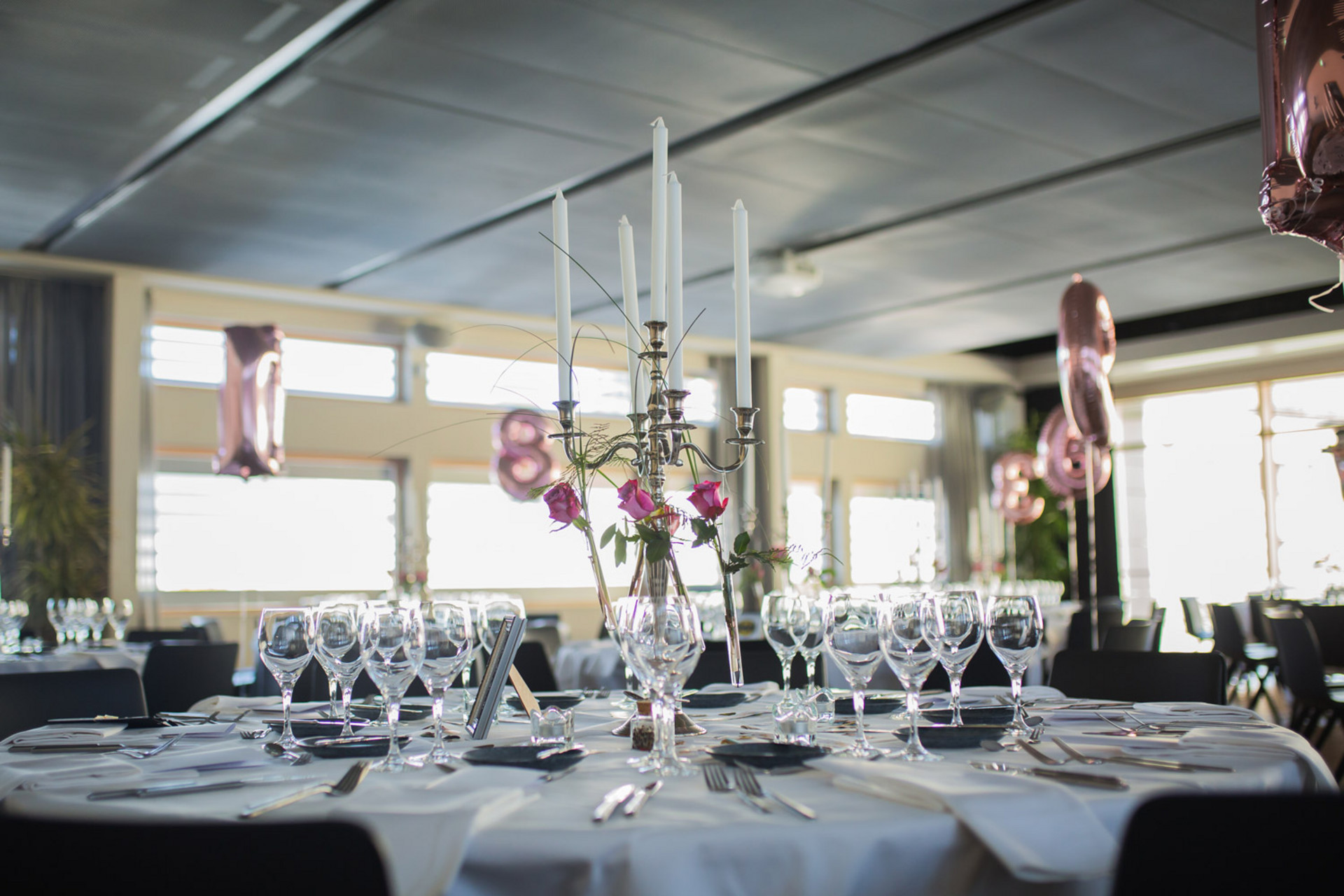 Eleganter runder Hochzeitstisch mit weißer Tischdecke, Gläsern, Besteck und hohem Kerzenleuchter mit rosa Blumen.