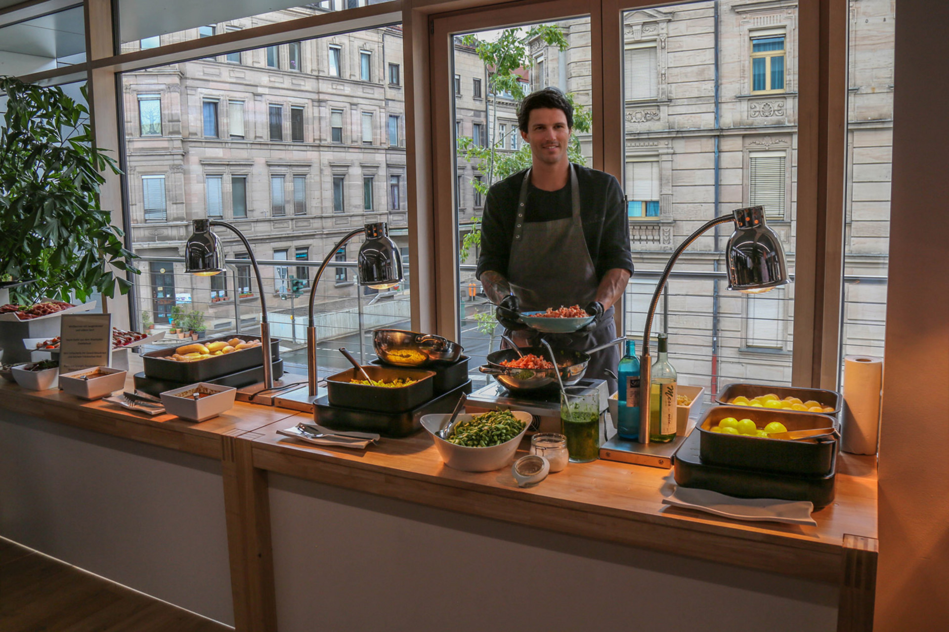 Buffetstation mit verschiedenen Gerichten, frischem Grün und Beilagen auf einer Holztheke vor großen Fenstern mit Blick auf ein historisches Fürther Gebäude.